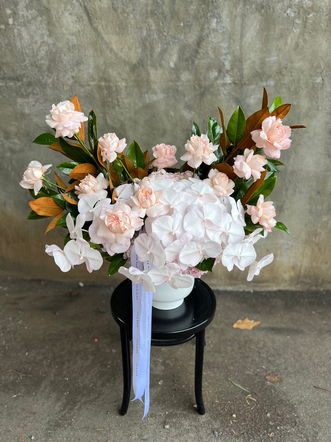 Large and luxe white ceramic vase displaying luxe blush pink flowers and magnolia foliage. Design sitting on a black bentwood chair with concrete wall behind.