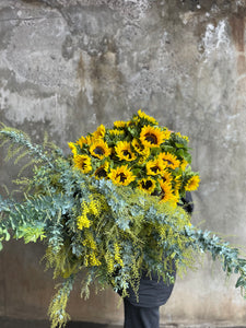 A large bundle of yellow sunflowers and yellow wattle held by a florist, wearing black and standing in front of a concrete wall.