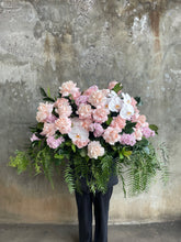 Wide image of Classic blush roses, blush hydrangeas, lilac stock and foliages feature in a Pastel pink, white and green casket design, held by a florist wearing back, up against a concrete wall.
