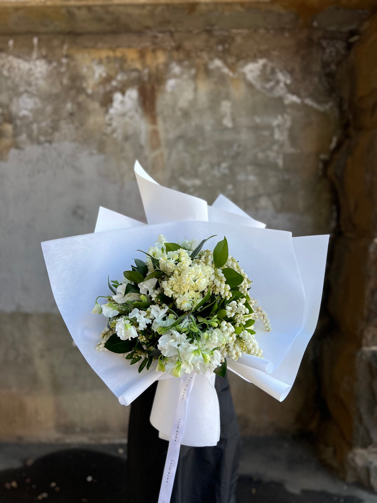 Florist holding September white and green spring bouquet, standing in front of concrete wall.
