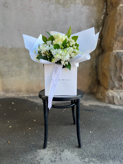 A white and green spring bouquet, presented in our Kate Hill Flower Bag. Bouquet in bag sitting on black bentwood chair in front of concrete wall.