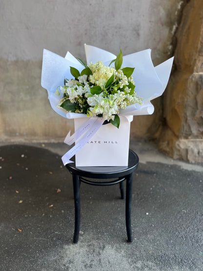 A white and green spring bouquet, presented in our Kate Hill Flower Bag. Bouquet in bag sitting on black bentwood chair in front of concrete wall.