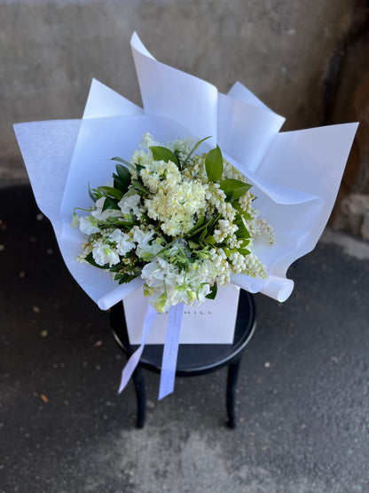 Image from above of white and green bouquet in flower bag. A white and green spring bouquet, presented in our Kate Hill Flower Bag. Bouquet in bag sitting on black bentwood chair in front of concrete wall.
