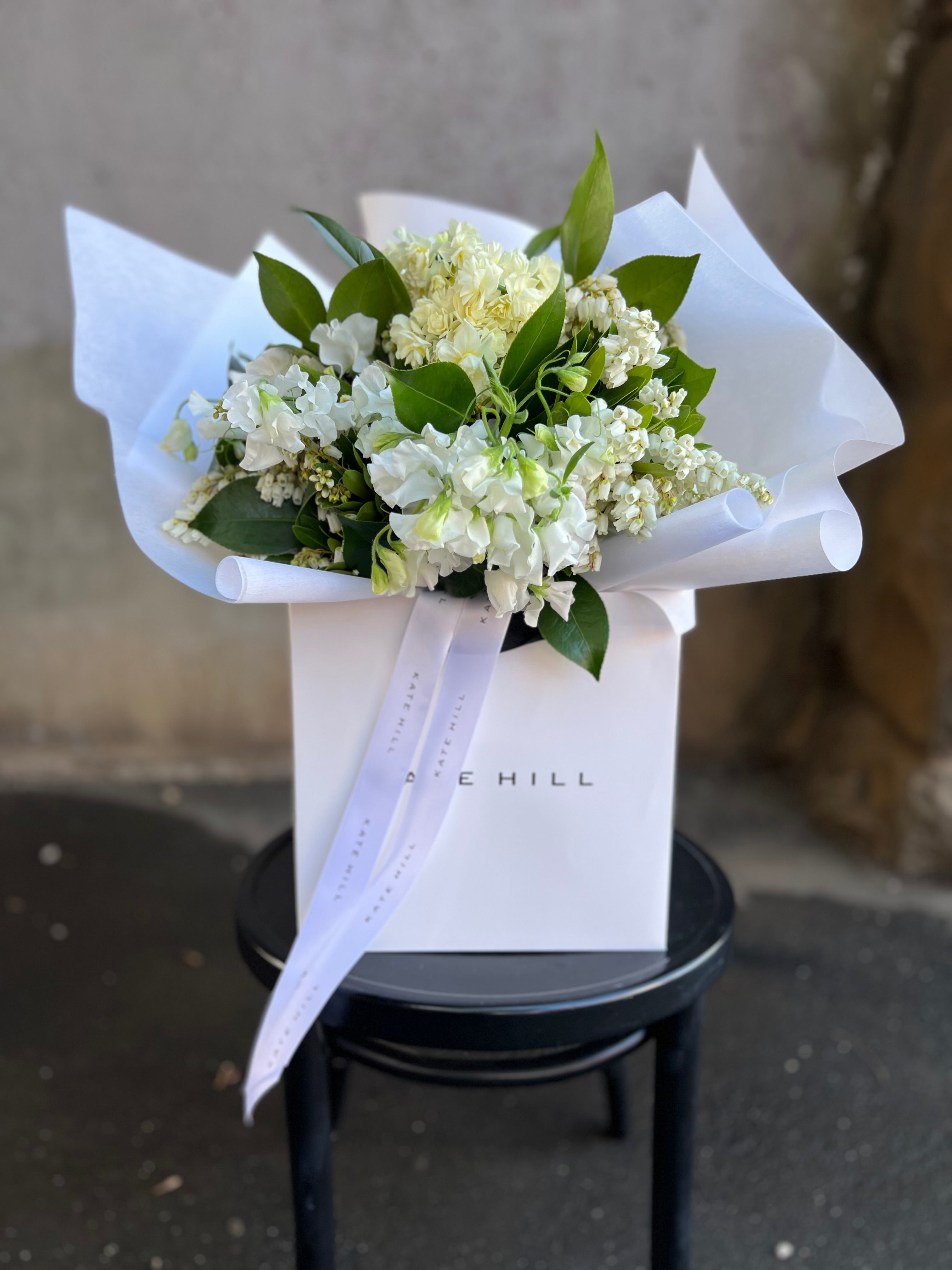 Front on image of white and green birthday bouquet. A white and green spring bouquet, presented in our Kate Hill Flower Bag. Bouquet in bag sitting on black bentwood chair in front of concrete wall.
