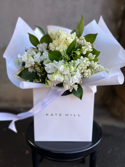 A white and green spring bouquet, presented in our Kate Hill Flower Bag. Bouquet in bag sitting on black bentwood chair in front of concrete wall.