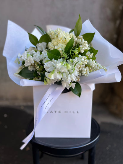 Up close image of white and green bouquet. A white and green spring bouquet, presented in our Kate Hill Flower Bag. Bouquet in bag sitting on black bentwood chair in front of concrete wall.
