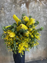 A large bundle of yellow native banksias and flowering yellow wattle, held by a florist, wearing black and standing in front of a concrete wall.