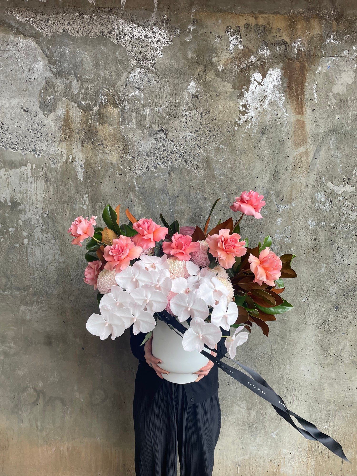 Large and luxe white ceramic vase displaying luxe watermelon roses, blush pink flowers and magnolia foliage. Design sitting on a black bentwood chair with concrete wall behind.