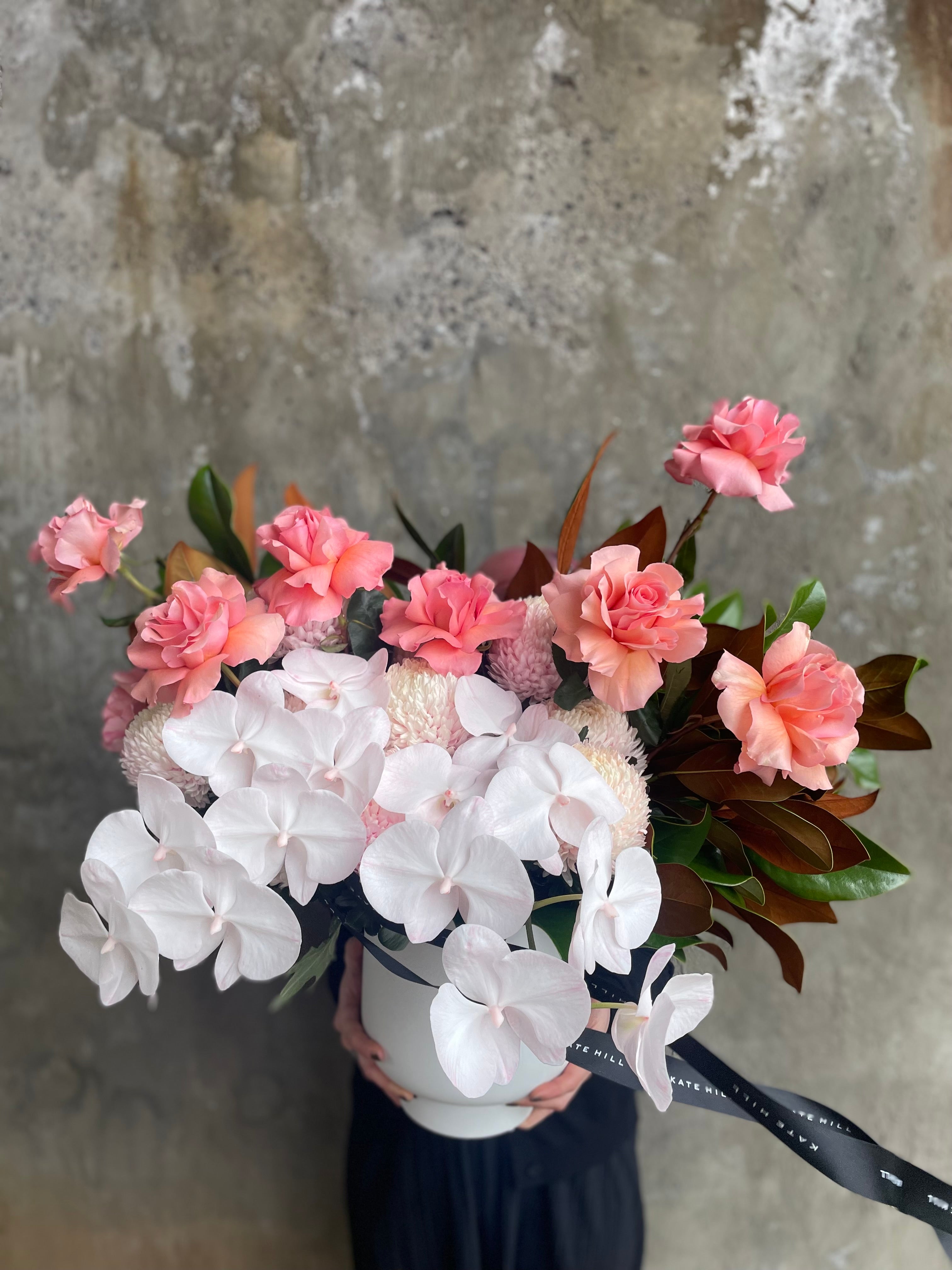 Close up image of large and luxe white ceramic vase displaying luxe blush pink flowers and magnolia foliage. Design sitting on a black bentwood chair with concrete wall behind.