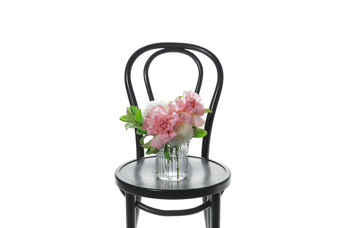 A mixed white, pink and green flower petite vase design sitting on a black bentwood chair with a white wall background.