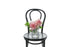 A mixed white, pink and green flower petite vase design sitting on a black bentwood chair with a white wall background.