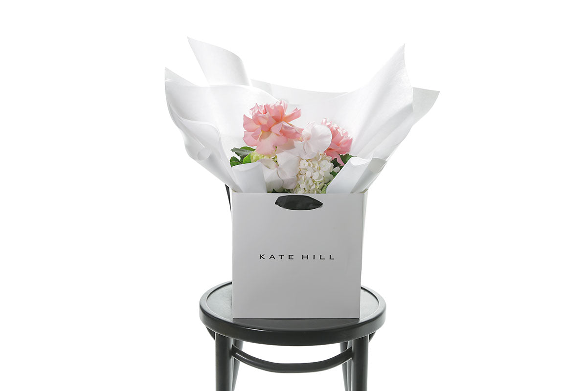 A mixed pastel flower bouquet beautifully wrapped in Kate Hill Flowers signature wrapping in our flower bag and sitting on a black bentwood chair with white wall background.