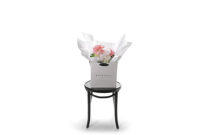 Wide image of a mixed pastel flower bouquet beautifully wrapped in Kate Hill Flowers signature wrapping in our flower bag and sitting on a black bentwood chair with white wall background.