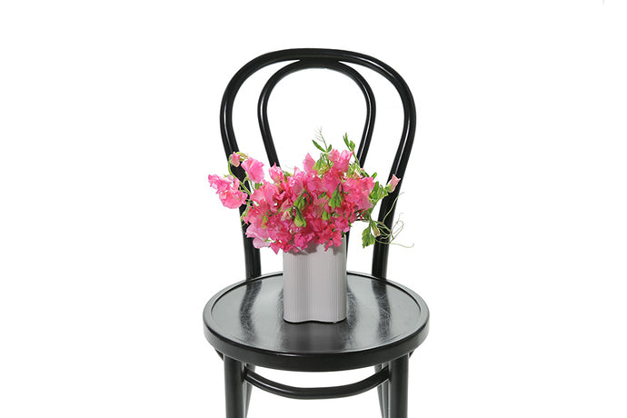 A miniature white wave vase displaying a small posy of vibrant pink sweetpea. Vase sitting on a black bentwood chair with white background.
