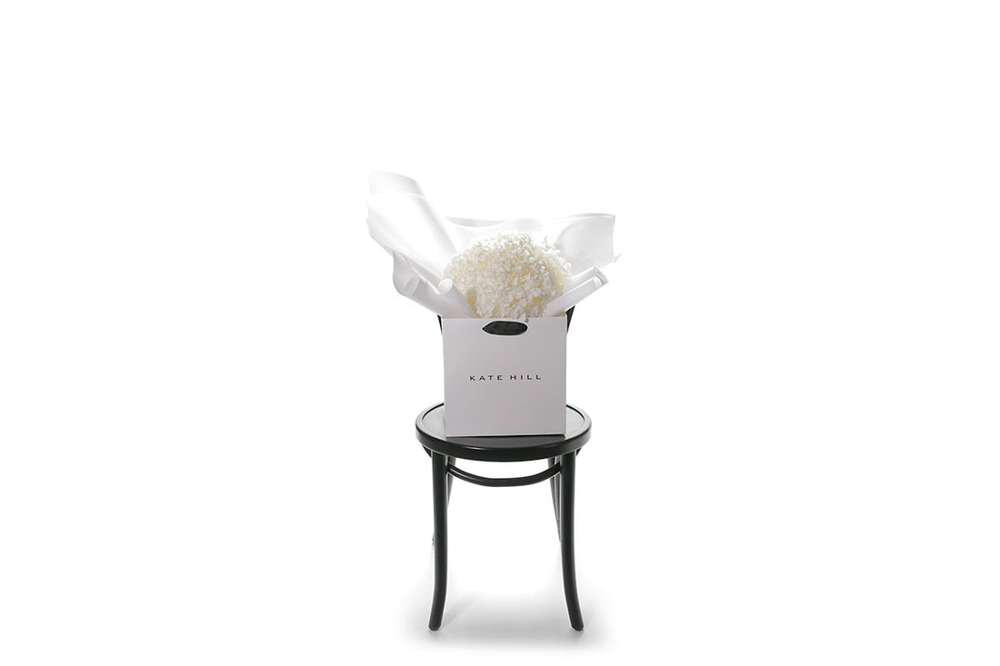 A bouquet of white bouquet of preserved dried hydrangeas, beautifully presented in signature white wrapping and flower bag. Flower bouquet sitting on a black bentwood chair with white wall background.