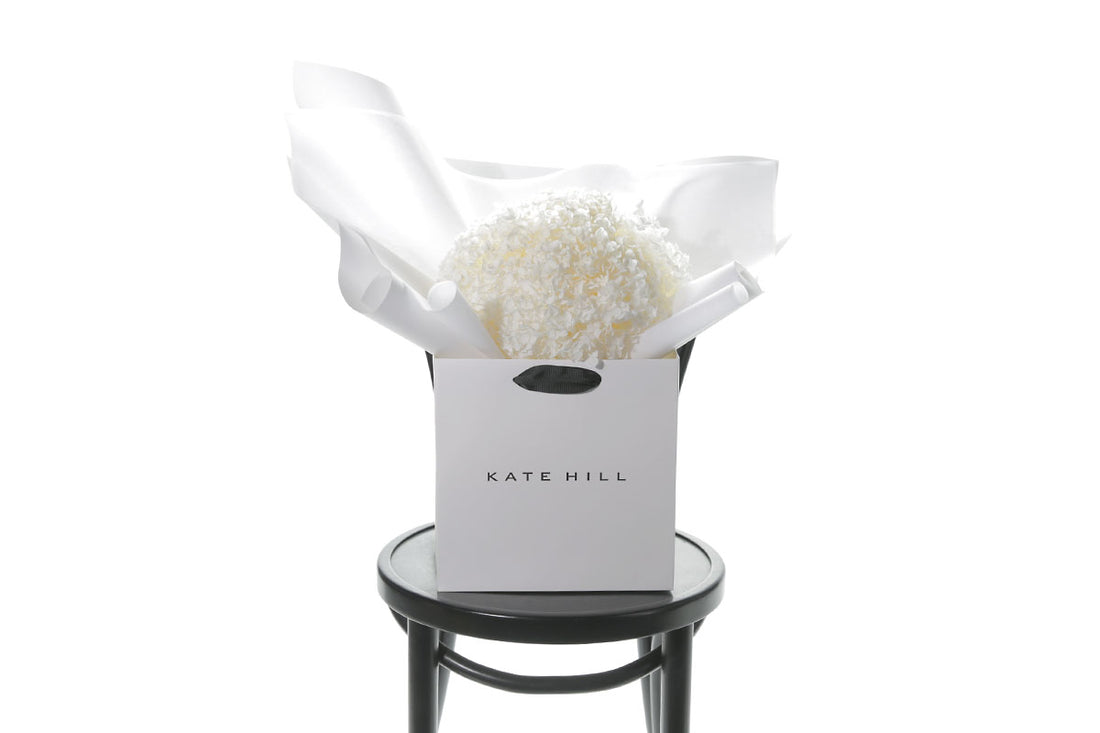 Close up image of a bouquet of candy pink bouquet of preserved dried hydrangeas, beautifully presented in signature white wrapping and flower bag. Flower bouquet sitting on a black bentwood chair with white wall background.