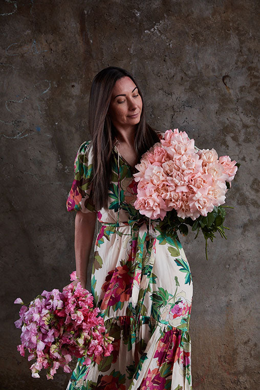 Florist Kate Hill holding bunches of flowers in a floral dress with a concrete backdrop