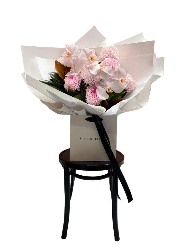 Wide image of large flower bouquet of blush pink flowers, beautifully wrapped in signature white paper and sitting in Kate Hill flower bag on black bentwood chair.