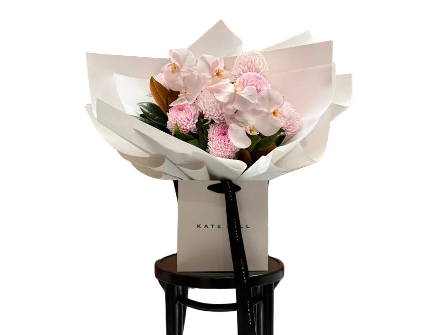 Close up image of large flower bouquet of blush pink flowers, beautifully wrapped in signature white paper and sitting in Kate Hill flower bag on black bentwood chair.