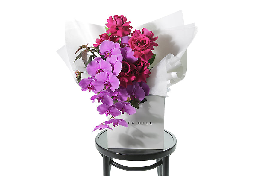 Kate Hill Flowers – large pink orchid and hot-pink rose graduation bouquet, hand-tied on a black chair with a white background.