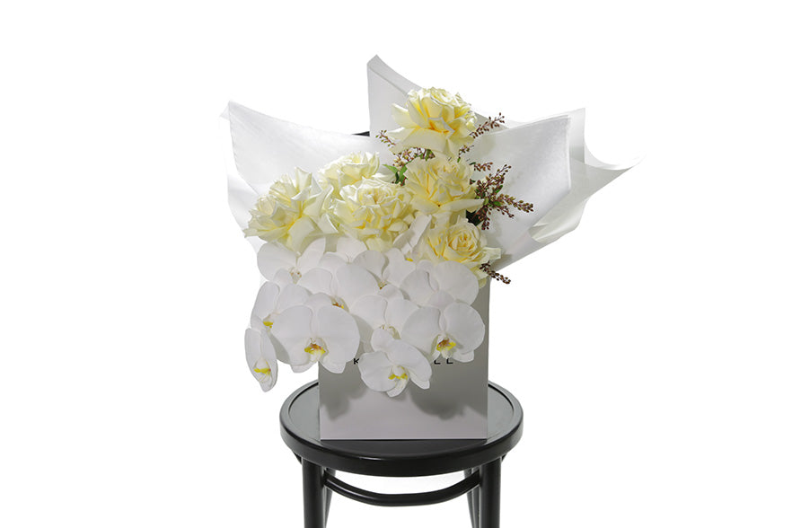 A large pastel bouquet named Lola. White or lemon butter roses, phalaenopsis orchid stems and seasonal foliage large bouquet. Flower bouquet is wrapped in signature white wrapping and in Kate Hill Flower bag. Bouquet bag sitting on a black bentwood chair with white background.