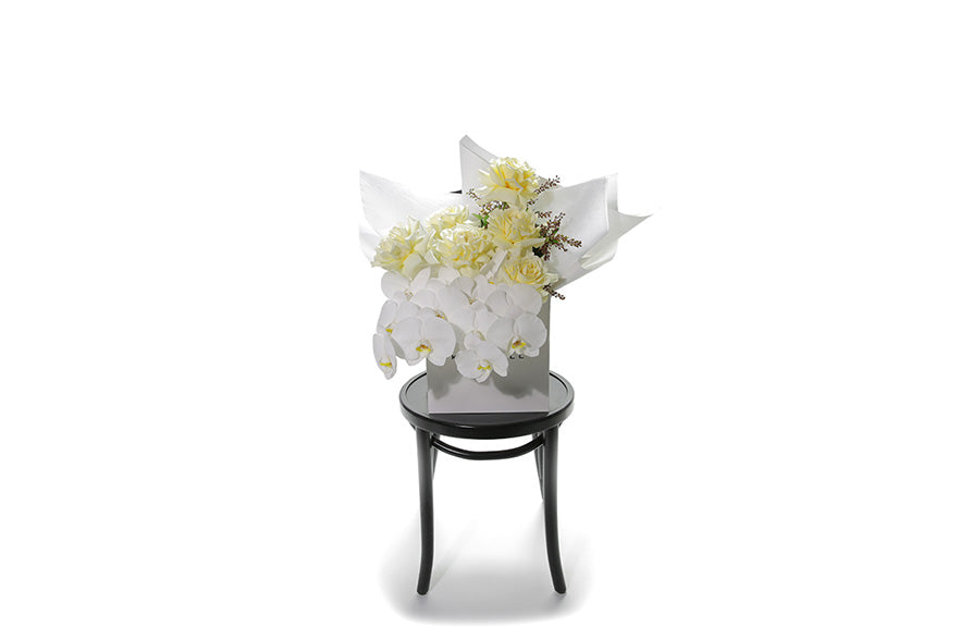 Wide image of Lola Bouquet. A large pastel bouquet named Lola. White or lemon butter roses, phalaenopsis orchid stems and seasonal foliage large bouquet. Flower bouquet is wrapped in signature white wrapping and in Kate Hill Flower bag. Bouquet bag sitting on a black bentwood chair with white background.
