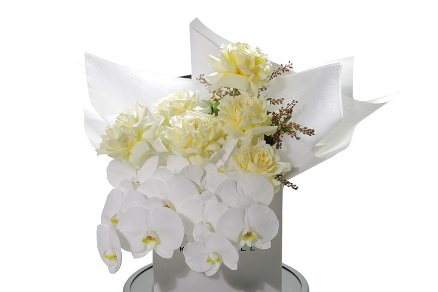 Close up image of Layla bouquet. A large pastel bouquet named Lola. White or lemon butter roses, phalaenopsis orchid stems and seasonal foliage large bouquet. Flower bouquet is wrapped in signature white wrapping and in Kate Hill Flower bag. Bouquet bag sitting on a black bentwood chair with white background.