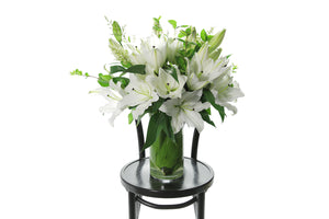 Open white lilies and green foliage displayed in a tapered vase lined with green leaves. Vase design sitting on black bentwood chair with white background.