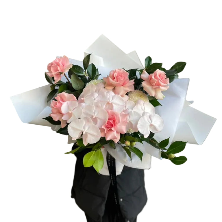 A florist in black holding a large May Birthday Bouquet. Large Bouquet f flowers in soft blush tones, creams and green foliages. Bouquet wrapped in our signature white presentation.
