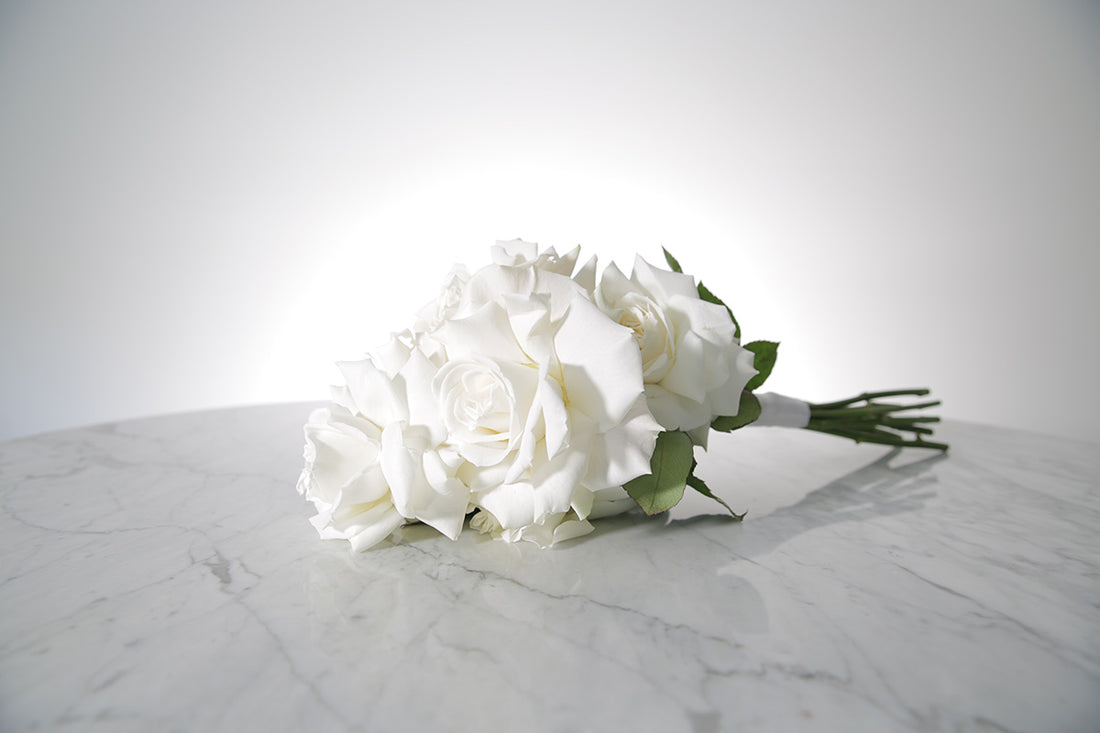 Reflexed white rose bridesmaid bouquet finished with ivory satin ribbon and pearl pin on a marble surface with a light grey background