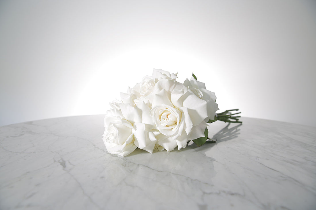 Reflexed white rose bridesmaid bouquet finished with ivory satin ribbon and pearl pin on a marble surface with a light grey background