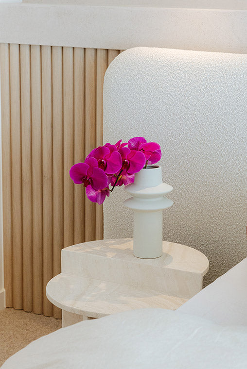 White vase with pink orchids on a marble nightstand against a textured wall.