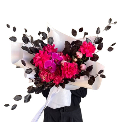 A florist holding a large and bright bouquet in bright pink tones, wrapped beautifully in white and standing against a white wall.