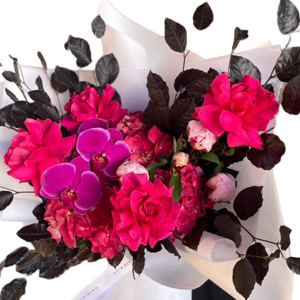 A close up of large and bright bouquet in bright pink tones, wrapped beautifully in white and standing against a white wall.