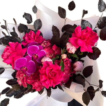 A close up of large and bright bouquet in bright pink tones, wrapped beautifully in white and standing against a white wall.