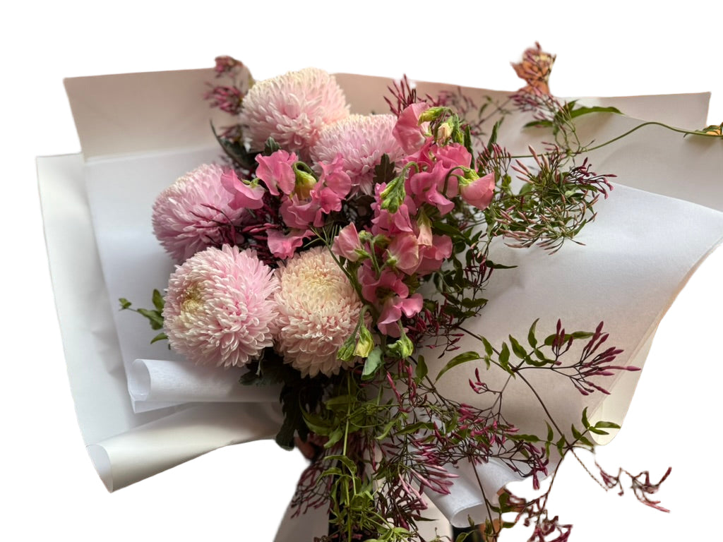Close p image of A soft pink flower bouquet of blush disbuds, sweet peas and jasmine foliage wrapped beautifully in signature white paper with Kate Hill ribbon. Florist holding bouquet against a white wall.