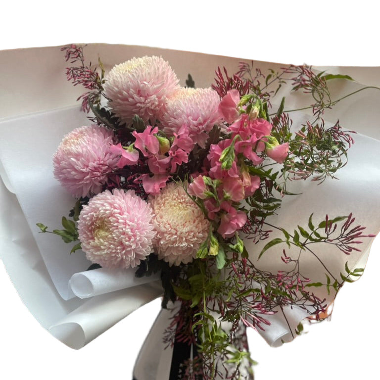 A soft pink flower bouquet of blush disbuds, sweet peas and jasmine foliage wrapped beautifully in signature white paper with Kate Hill ribbon. Florist holding bouquet against a white wall.