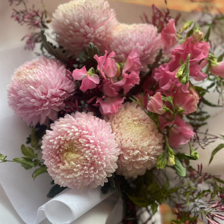Close up image of A soft pink flower bouquet of blush disbuds, sweet peas and jasmine foliage wrapped beautifully in signature white paper with Kate Hill ribbon. Florist holding bouquet against a white wall.