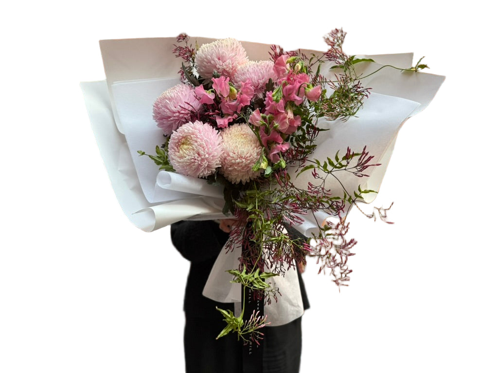 A soft pink flower bouquet of blush disbuds, sweet peas and jasmine foliage wrapped beautifully in signature white paper with Kate Hill ribbon. Florist holding bouquet against a white wall.
