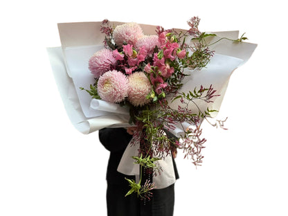 A soft pink flower bouquet of blush disbuds, sweet peas and jasmine foliage wrapped beautifully in signature white paper with Kate Hill ribbon. Florist holding bouquet against a white wall.