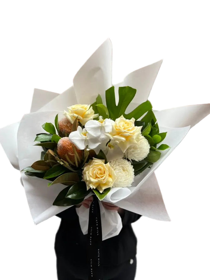 Florist holding our October Birthday Bouquet featuring lemon roses, banksias, white disbuds, white orchid and seasonal foliage. Presented beautifully in our signature white wrapping.