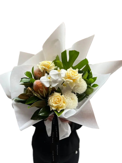 Florist holding our October Birthday Bouquet featuring lemon roses, banksias, white disbuds, white orchid and seasonal foliage. Presented beautifully in our signature white wrapping.