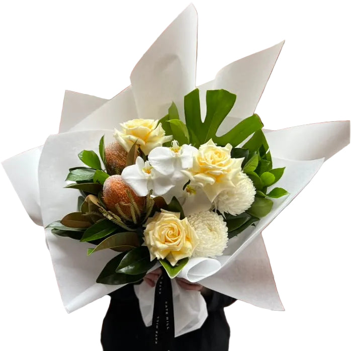 Florist holding our October Birthday Bouquet featuring lemon roses, banksias, white disbuds, white orchid and seasonal foliage. Presented beautifully in our signature white wrapping.