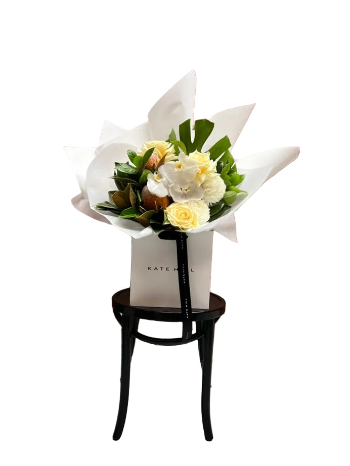 Kate Hill flower bag sitting on a black bentwood chair with our October Birthday Bouquet featuring lemon roses, banksias, white disbuds, white orchid and seasonal foliage. Presented beautifully in our signature white wrapping.