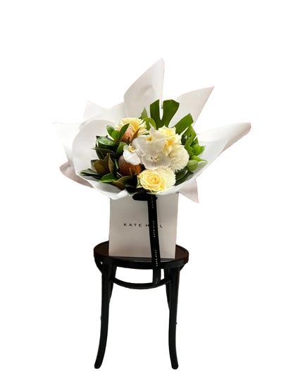 Kate Hill flower bag sitting on a black bentwood chair with our October Birthday Bouquet featuring lemon roses, banksias, white disbuds, white orchid and seasonal foliage. Presented beautifully in our signature white wrapping.