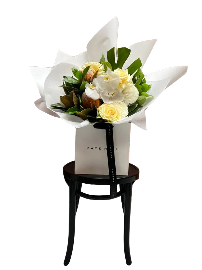 Kate Hill flower bag sitting on a black bentwood chair with our October Birthday Bouquet featuring lemon roses, banksias, white disbuds, white orchid and seasonal foliage. Presented beautifully in our signature white wrapping.