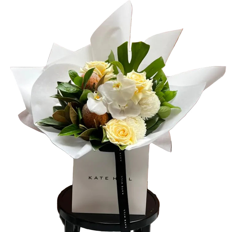 Kate Hill flower bag sitting on a black bentwood chair with a close up of our October Birthday Bouquet featuring lemon roses, banksias, white disbuds, white orchid and seasonal foliage. Presented beautifully in our signature white wrapping.