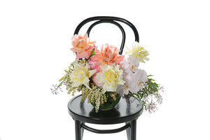 A 20cm ball vase, lined with green monsteria leaves, displaying soft pastel mixed flowers in ivory, lemon, blush and watermelon tones. Vase design sitting on a black bentwood chair with white background.