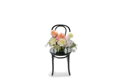 Wide image of Pascale vase design. A 20cm ball vase, lined with green monsteria leaves, displaying soft pastel mixed flowers in ivory, lemon, blush and watermelon tones. Vase design sitting on a black bentwood chair with white background.