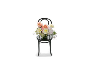 Wide image of Pascale vase design. A 20cm ball vase, lined with green monsteria leaves, displaying soft pastel mixed flowers in ivory, lemon, blush and watermelon tones. Vase design sitting on a black bentwood chair with white background.